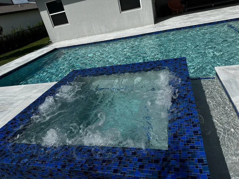 Raised spa with deep blue mosaic tile bubbling beside a pool and a gray house