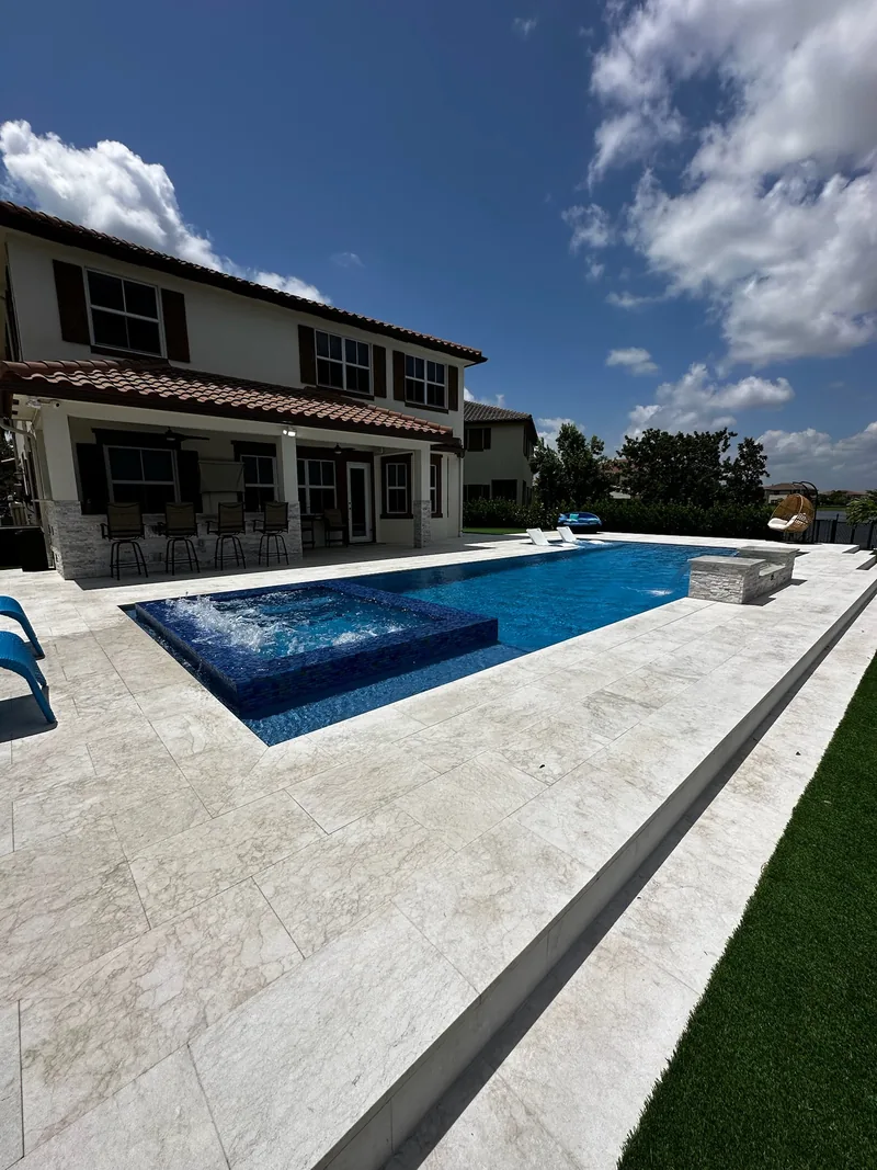 Blue pool with raised spa, light stone deck and a stepped turf edge beside a house