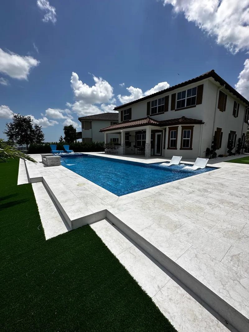 Blue pool with white loungers beside a two-story house and a green turf lawn