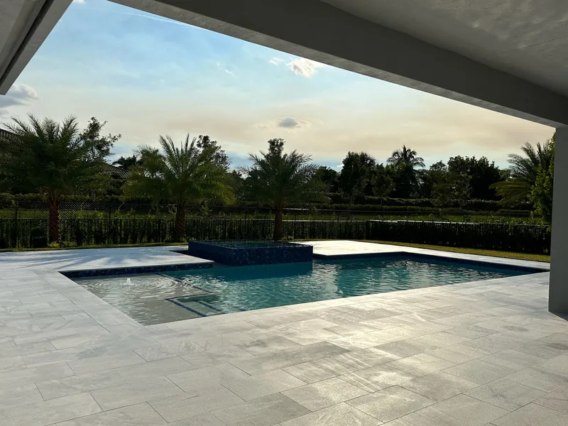 View from a covered patio over a pool with a raised spa and light paver deck at dusk