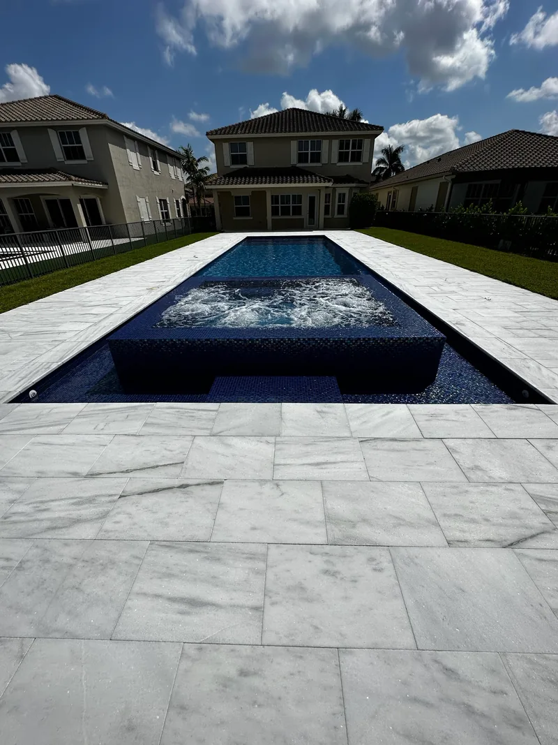 Bubbling raised spa with dark blue mosaic tile in a long pool and white veined paver deck