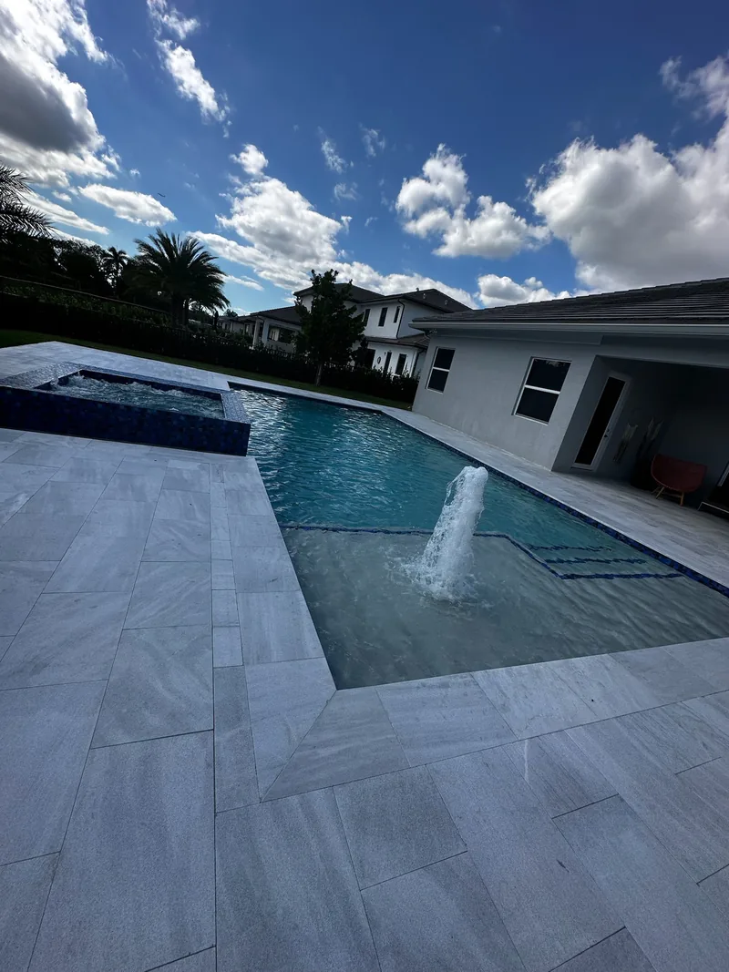 Deck jet water arc in a pool with light gray pavers, white house and blue sky