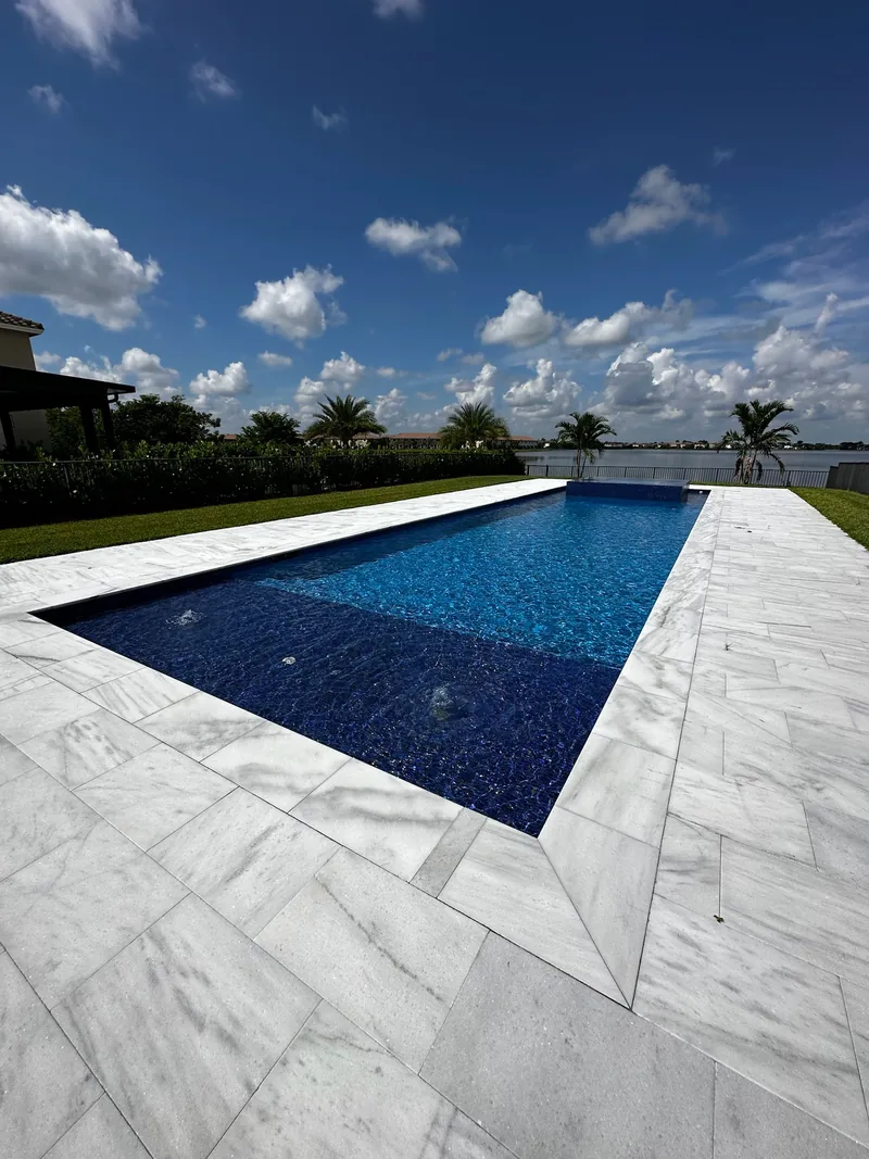 Deep blue mosaic pool with a white veined paver deck and lawn beside a lake