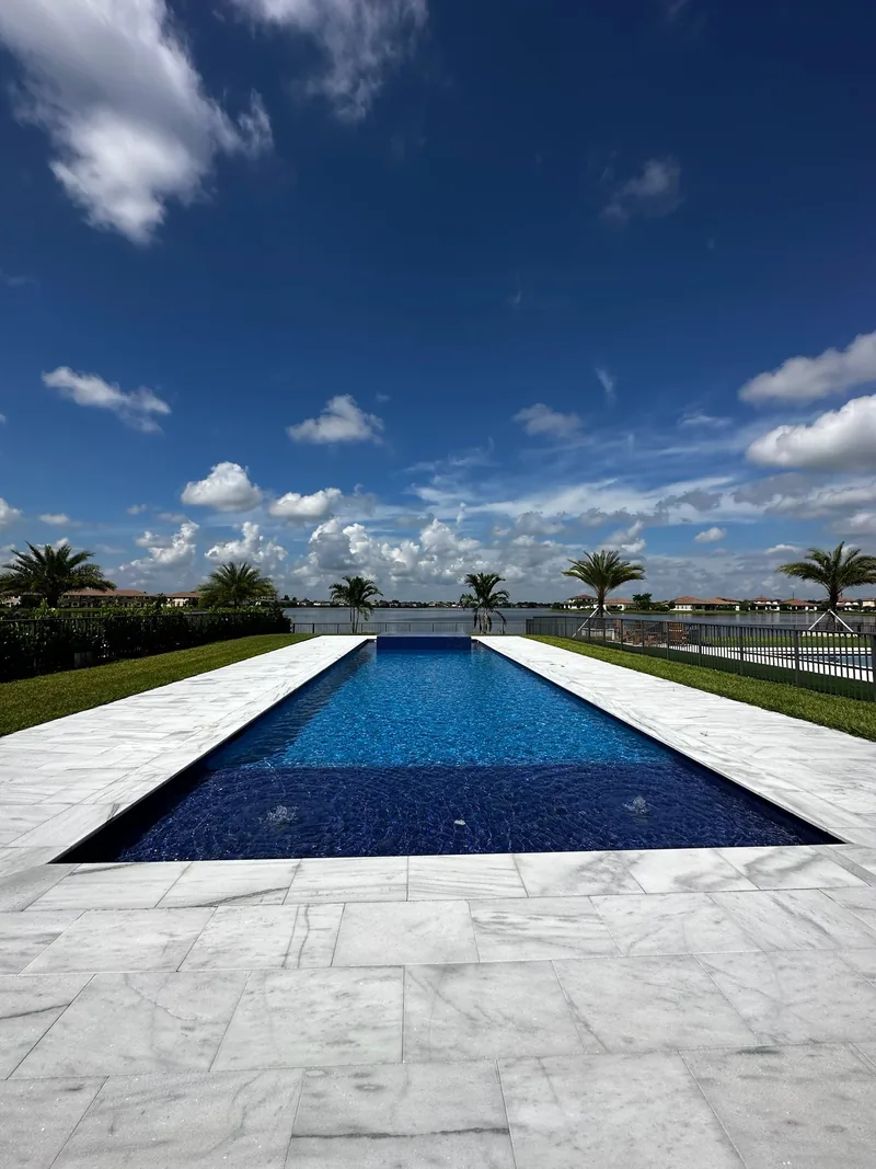 Infinity-edge pool with a deep blue mosaic floor and white veined paver deck in front of a lake