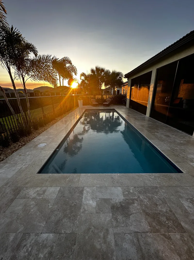 Long pool reflecting the sky at sunset next to a lanai and stone-style pavers