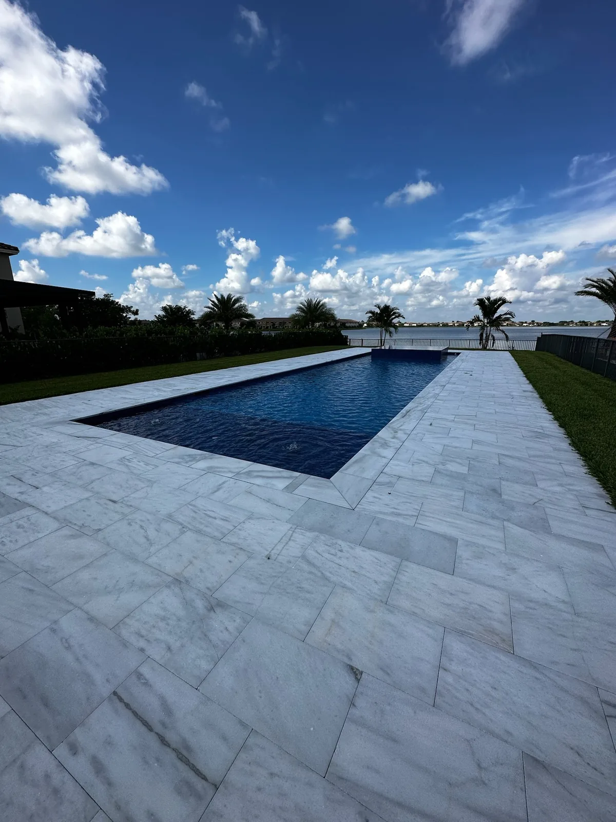 Long narrow pool with dark blue water and a wide white marble-style paver deck beside a lake