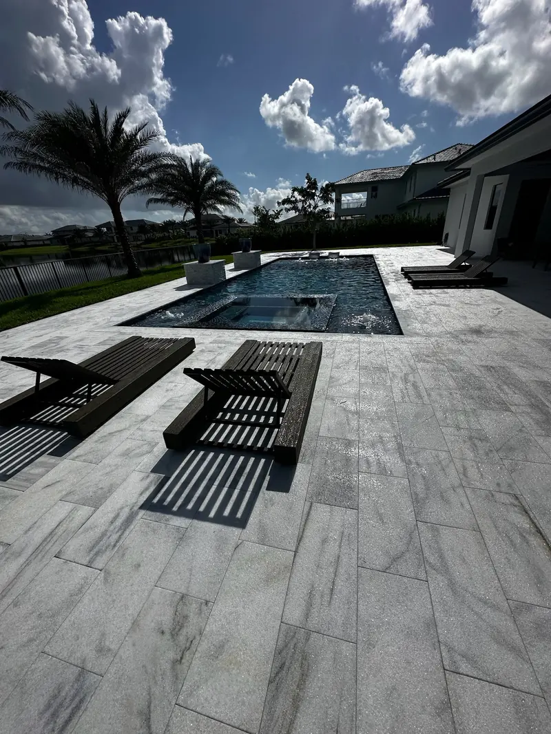 Light veined stone deck with black loungers and a pool with a spa overlooking a lake