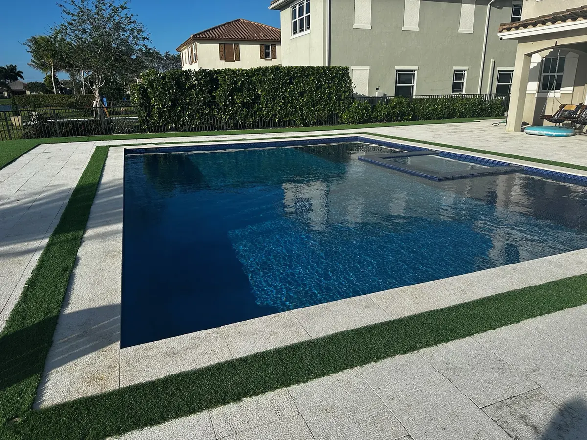 Pool corner with light stone coping, a turf strip and paver deck in front of a two-story house