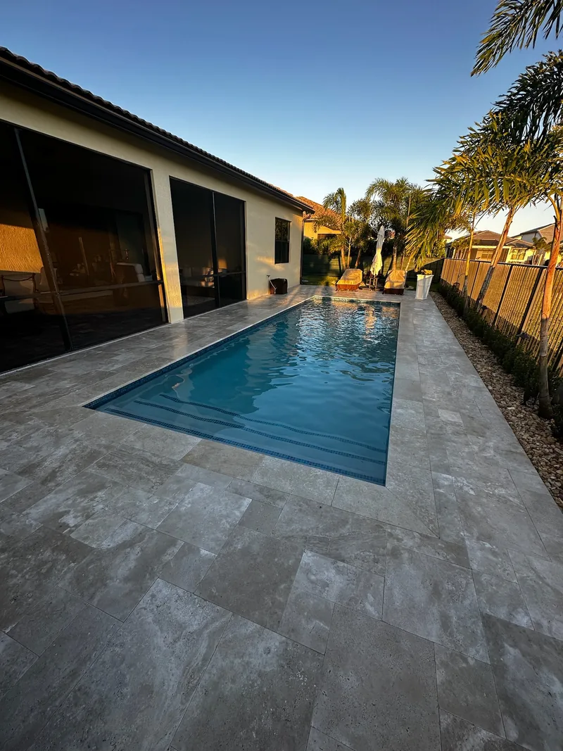 Narrow pool beside a screened lanai with stone-style pavers and a fence