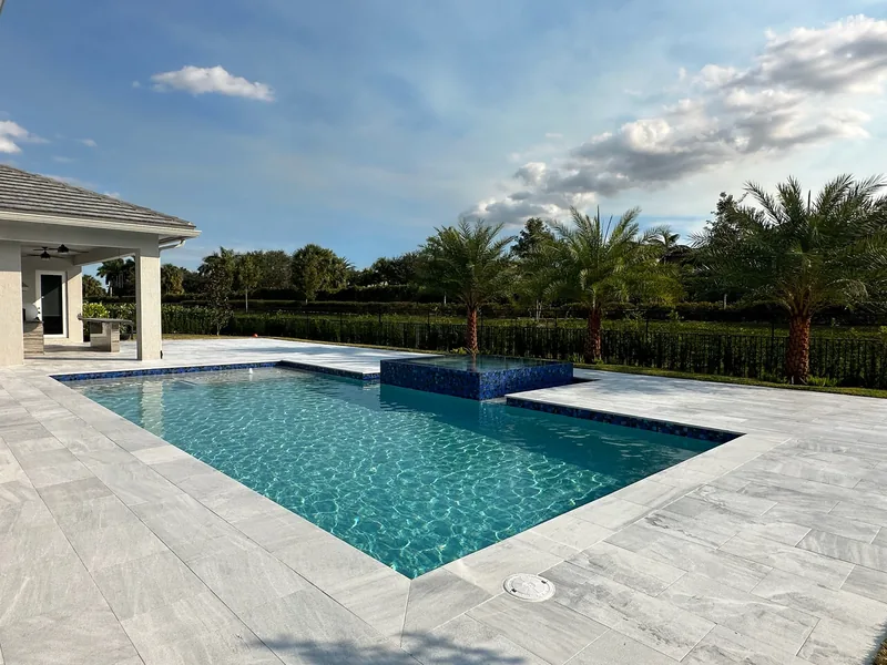 Turquoise pool with a mosaic raised spa and light gray pavers, with palms and hedge behind