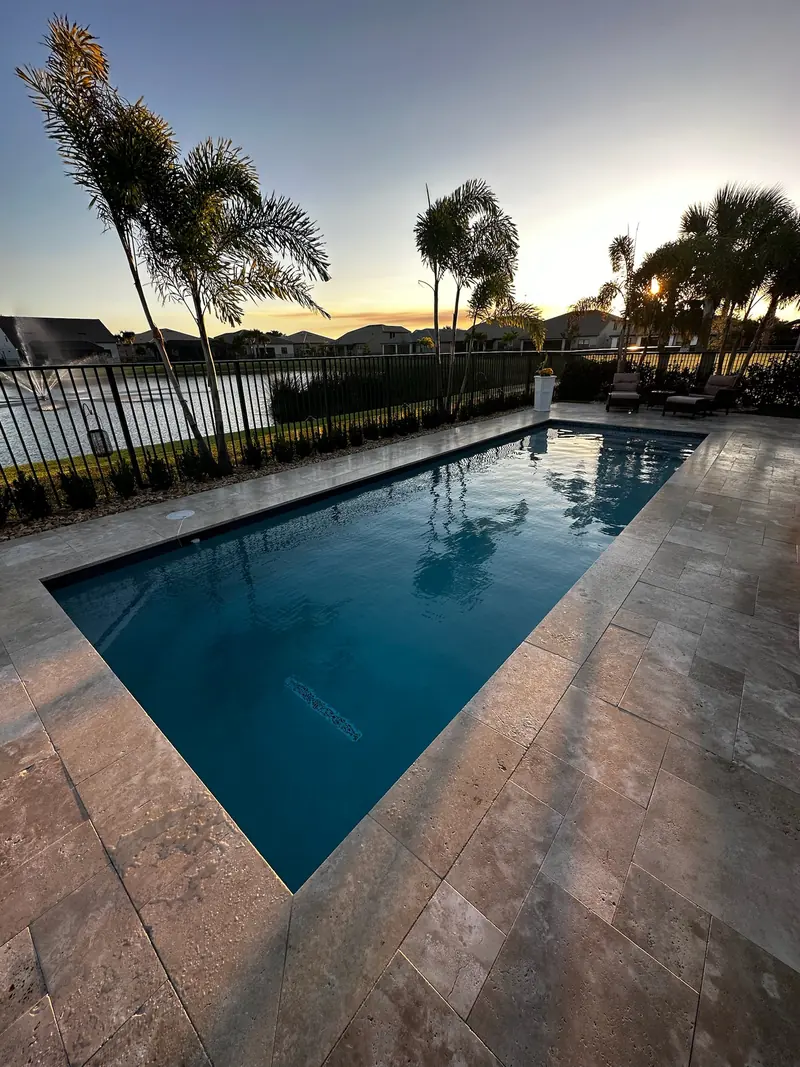 Pool reflecting sky and palms with a stone-style paver deck and a lake at sunset