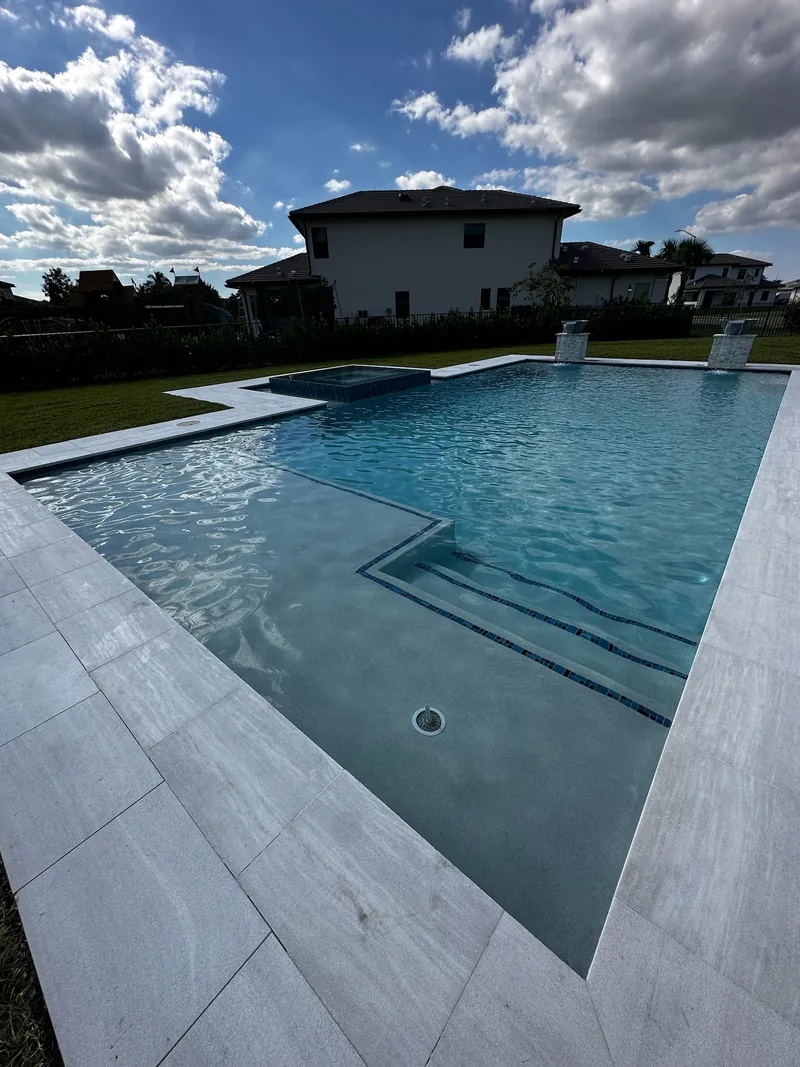Pool with entry steps and a shallow ledge, plank-style pavers and a two-story house behind
