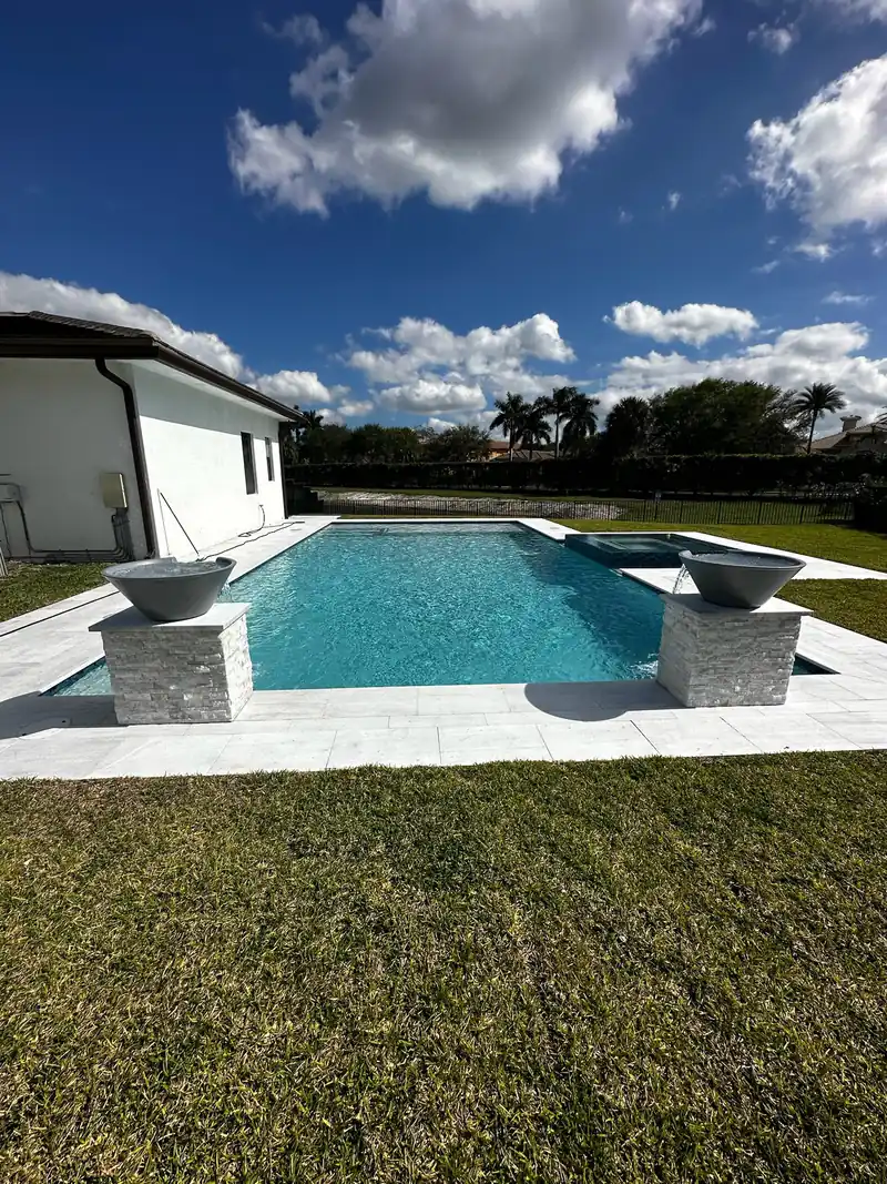 Pool with two stone-clad scupper bowls and light coping on a green lawn beside a white house