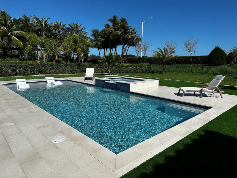 Turquoise pool with white coping, lounge chair and spa framed by palms and a hedge