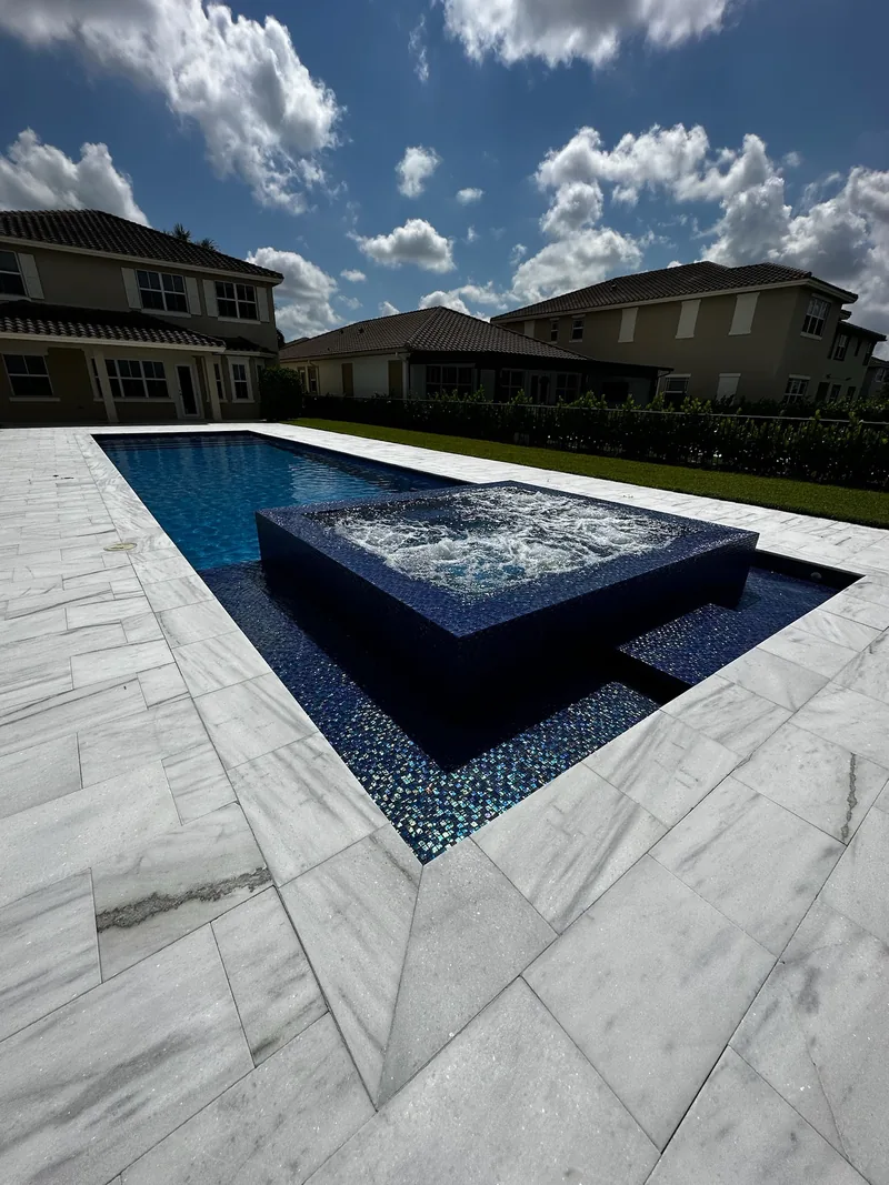 Corner view of a raised dark mosaic spa in a long pool with white veined pavers