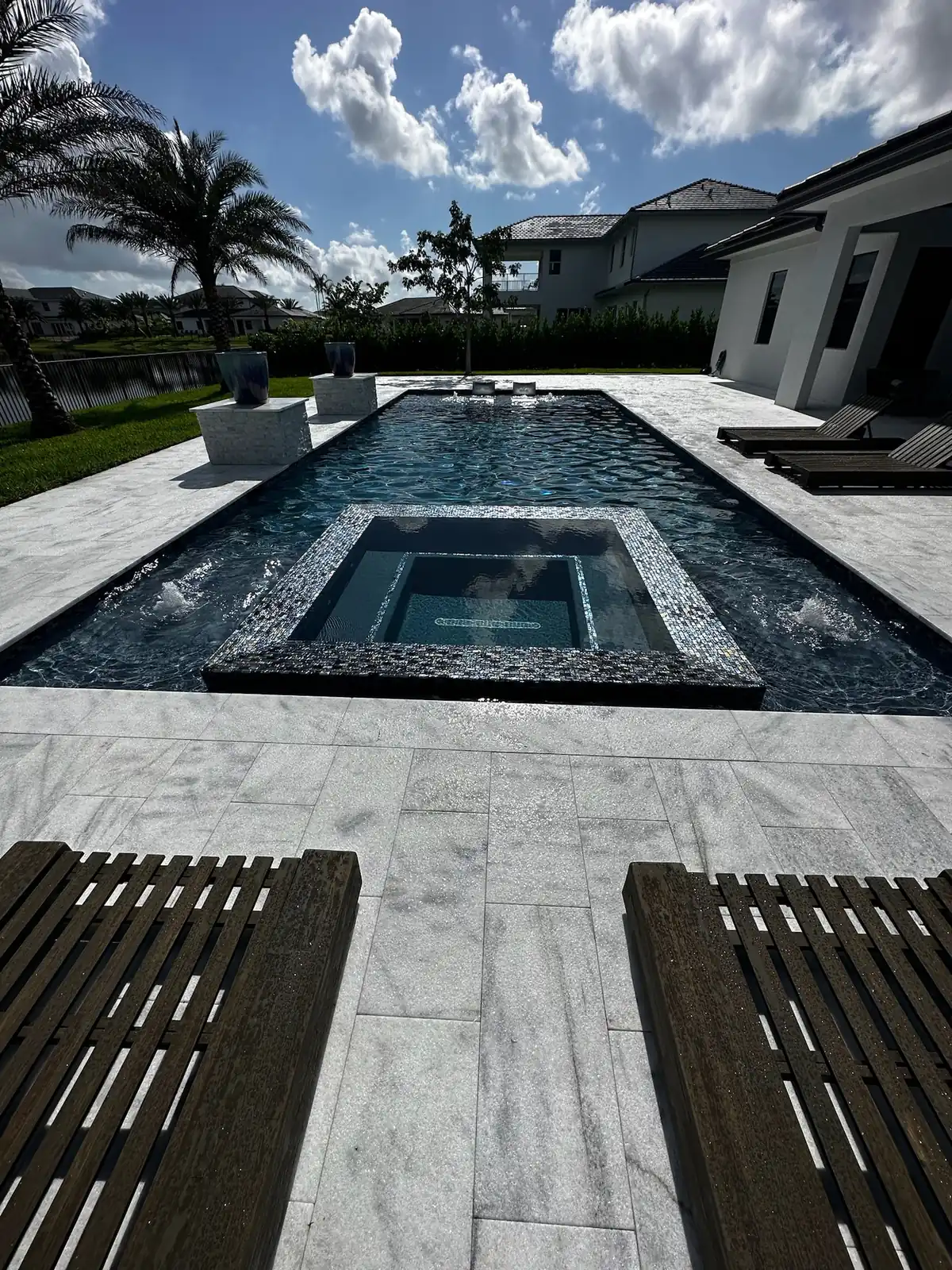 View between two dark loungers toward a spa and narrow pool on veined stone pavers