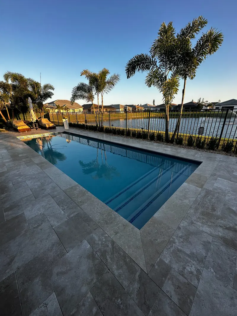 Rectangular pool with deep blue water, stone-style pavers and a lake with palms at sunset
