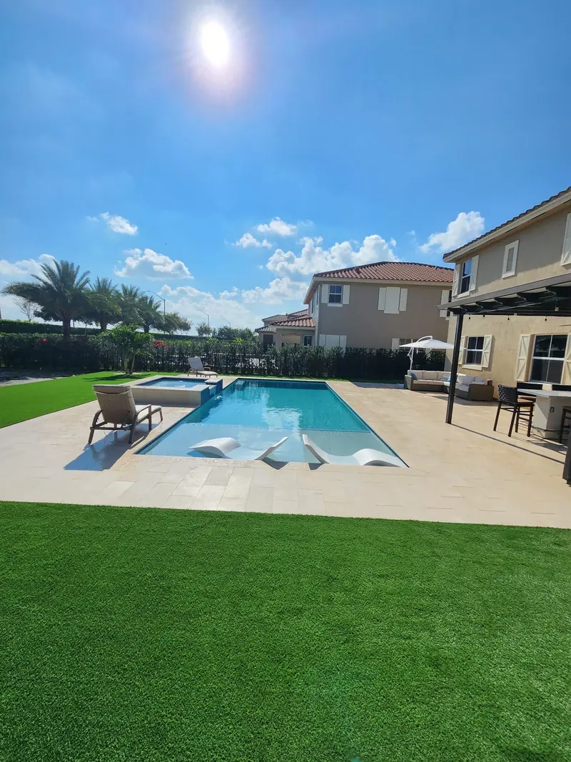 Pool with in-pool loungers and a light patio on a green turf lawn with a two-story house