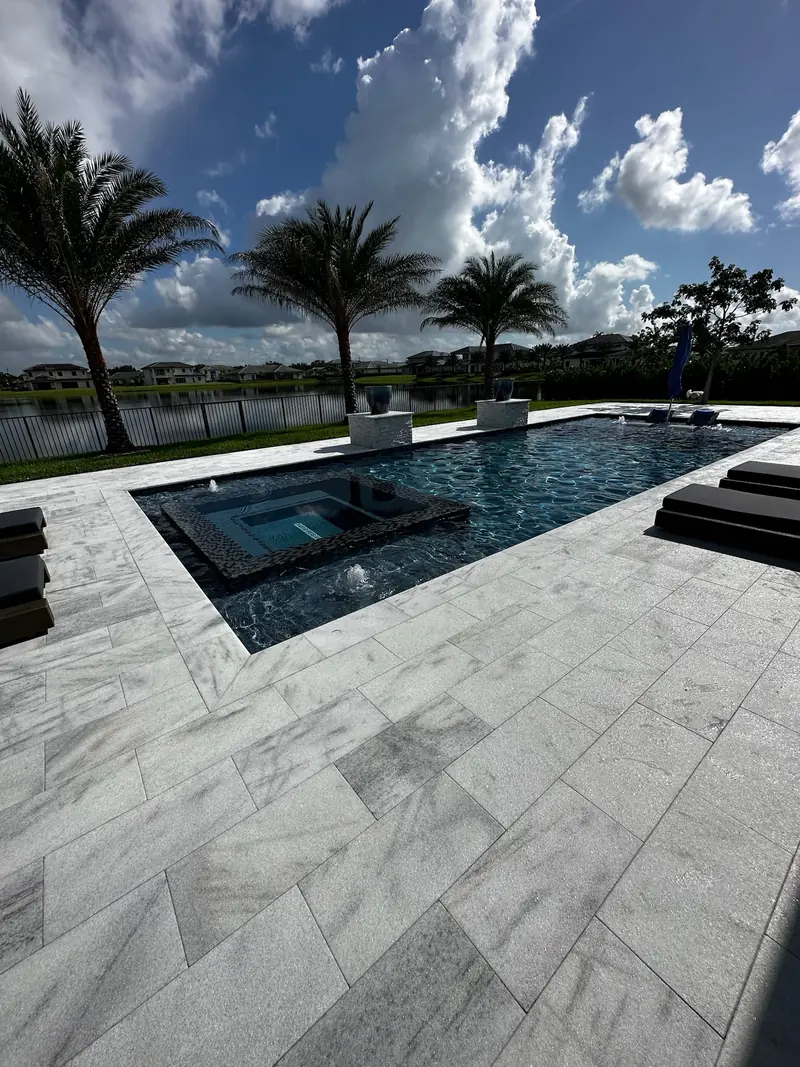 Veined stone deck leading to a pool with a dark mosaic spa, palms and a lake beyond