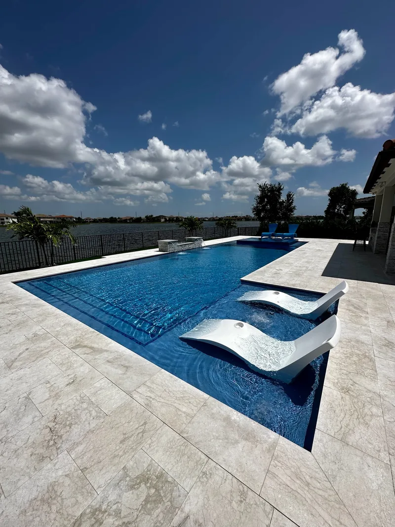 Vivid blue pool with white in-pool loungers on a travertine-style deck facing a lake