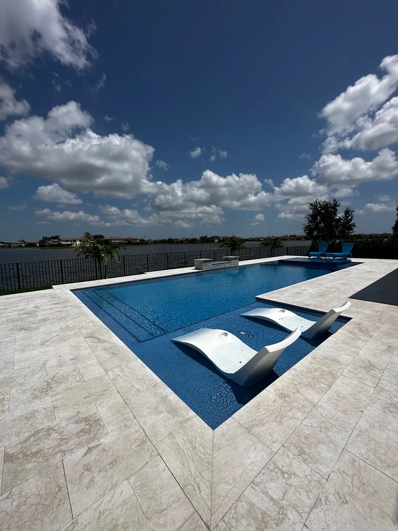 Two white in-pool loungers on a tanning ledge with a lake and sky behind the pool